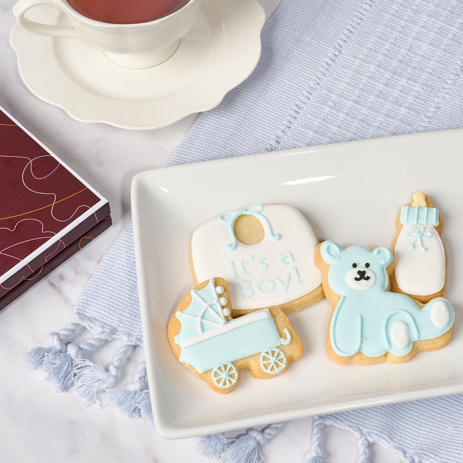 A set of decorated cookies designed in shapes of a baby rattle, a teddy bear, and a baby bottle, presented on a white plate with a cup and saucer in the background.